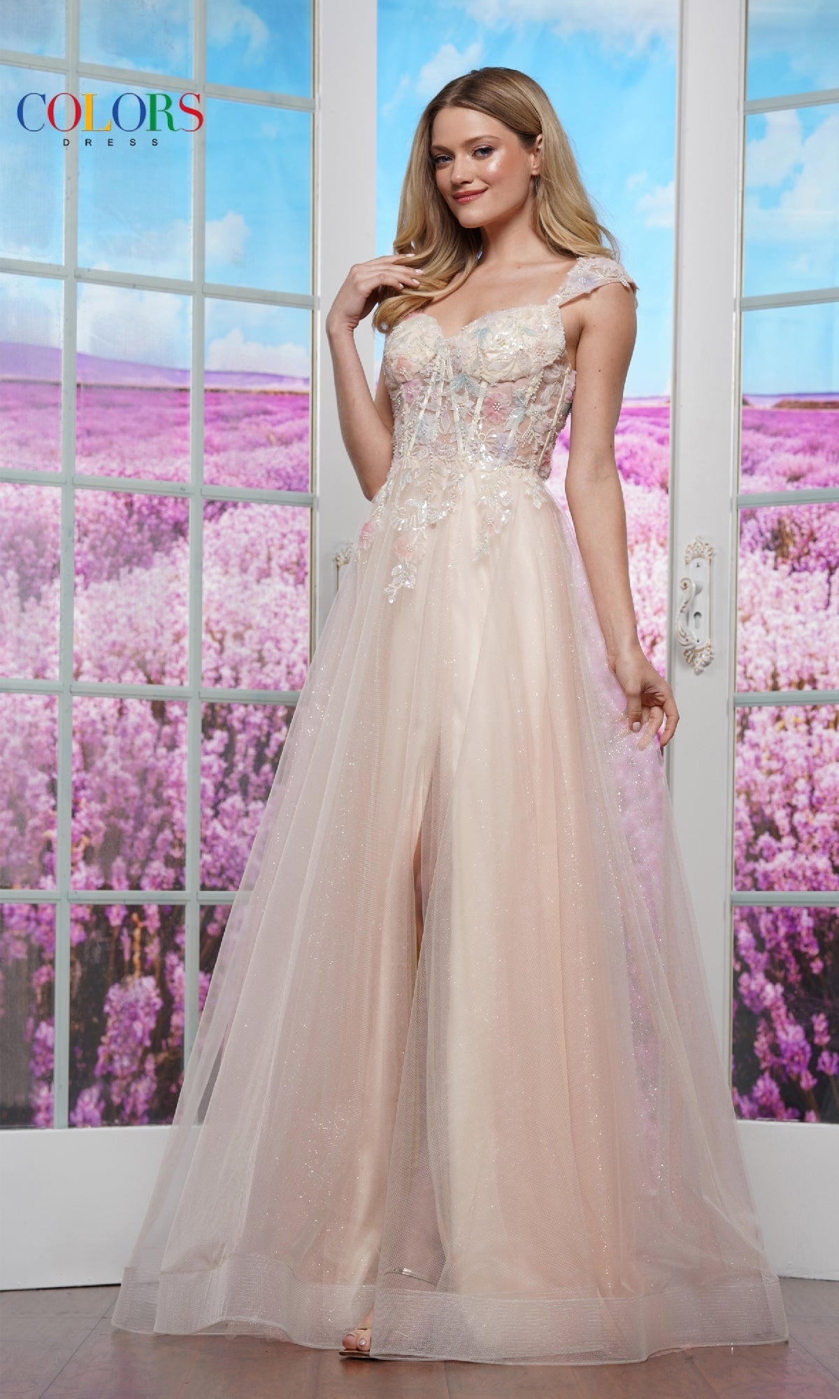 A woman wears the Colors Dress 3498, an off-shoulder glitter tulle prom ball gown in sheer light pink with a corset bodice, standing indoors before glass doors overlooking a purple flower field.